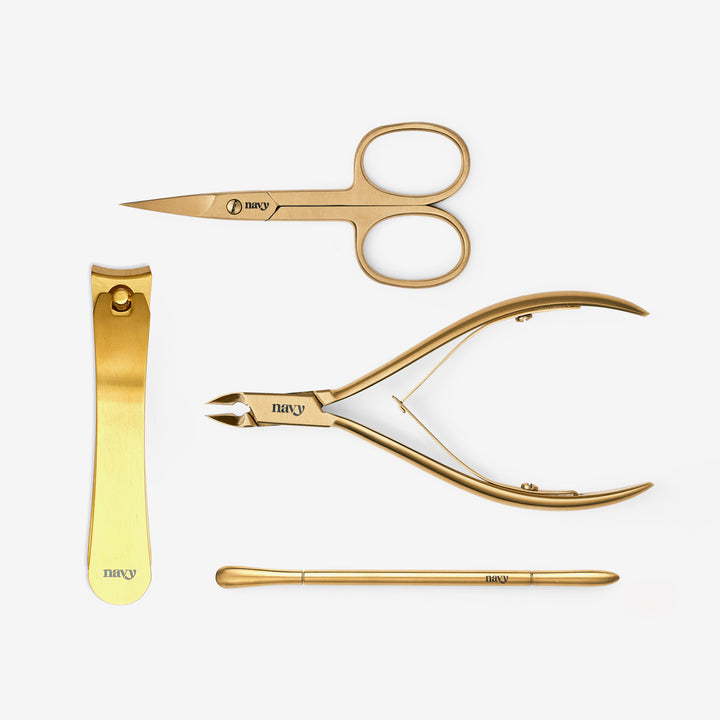 A collection of four gold-colored nail tools including toe nail nips, a curved pusher tool, fine cuticle nips, and curved scissors, all laid out on a white background.