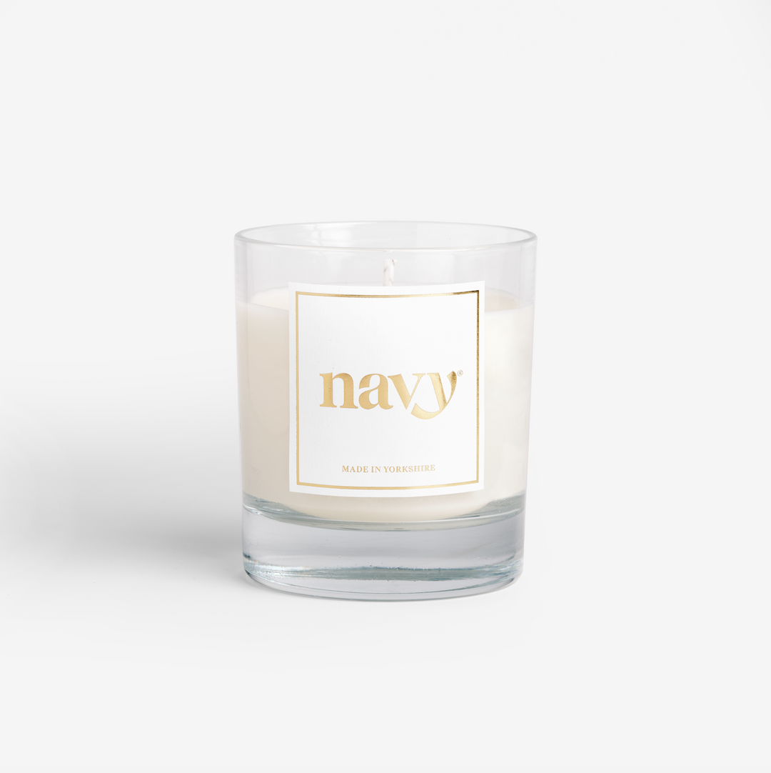 A white soy wax candle with a gold label that reads 'navy', set in a clear glass container. Signature scent