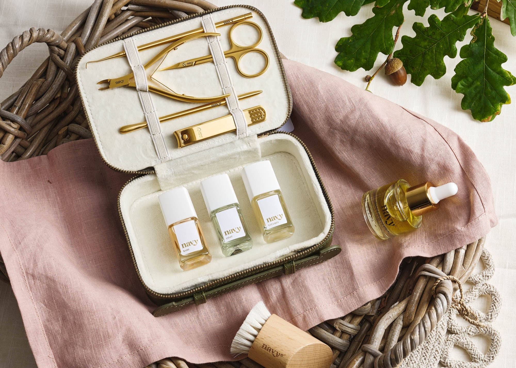 Cosmetic set with bottles, tools, and a brush on a pink cloth with green leaves in the background.