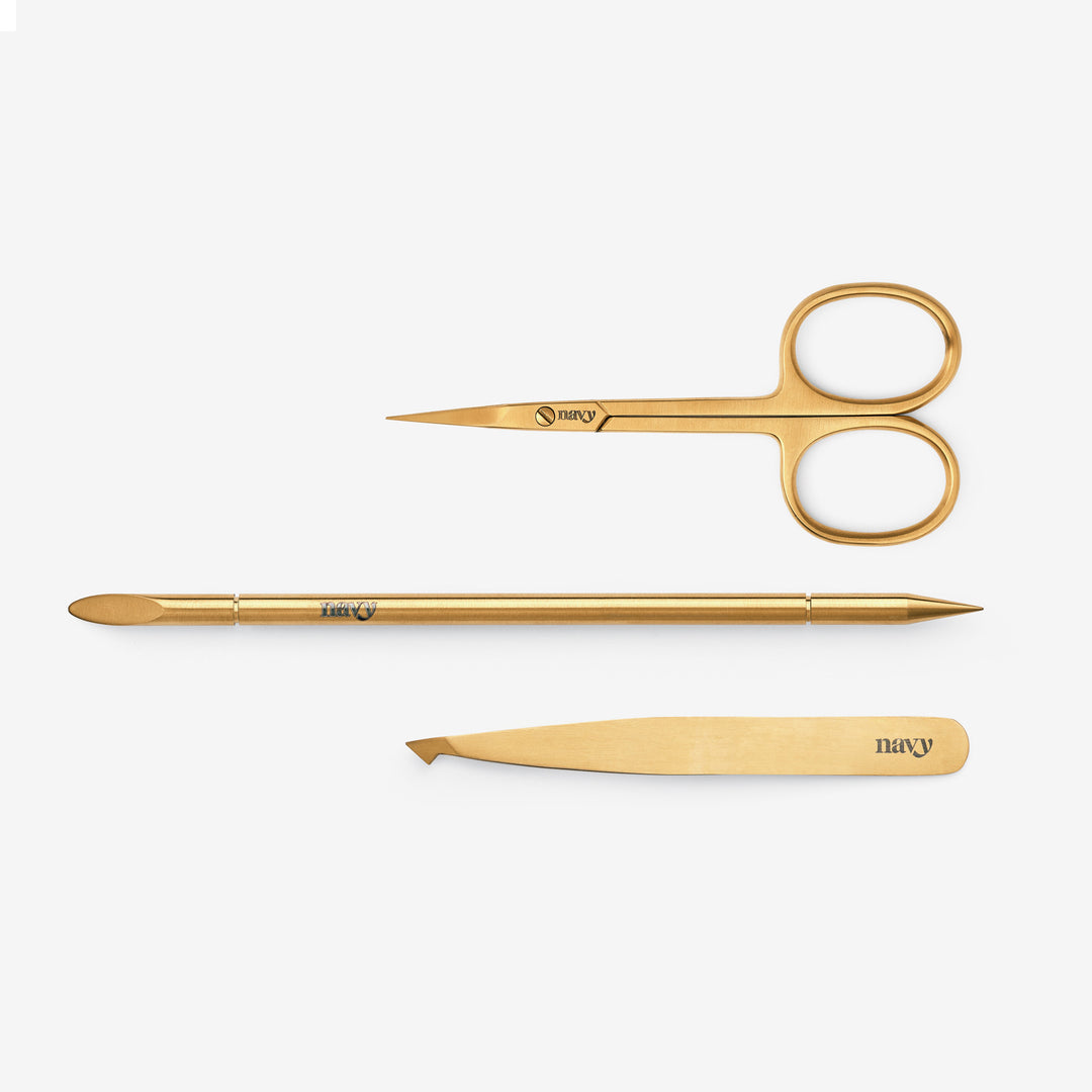 A collection of eyebrow grooming tools including a slant tweeze, straight scissor, and a reusable orange-wood stick.