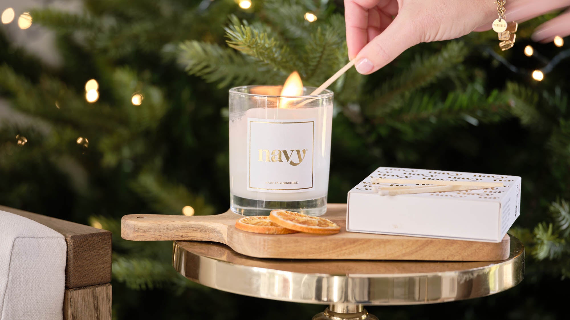Candle being lit with a match, surrounded by greenery and a box of matches on a small table.