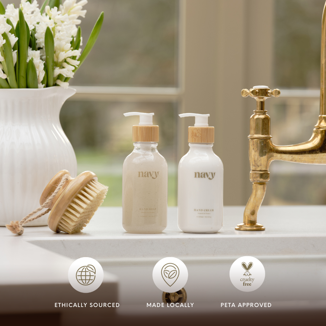 Navy Hand Care Duo next to gold taps on sink, a nail brush and a vase of flowers. 