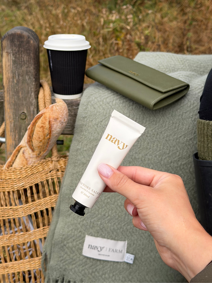 A hand holds a white tube of Navy Farm hand cream with gold lettering, set against an outdoor picnic scene with a coffee cup, bread, and an olive green blanket.