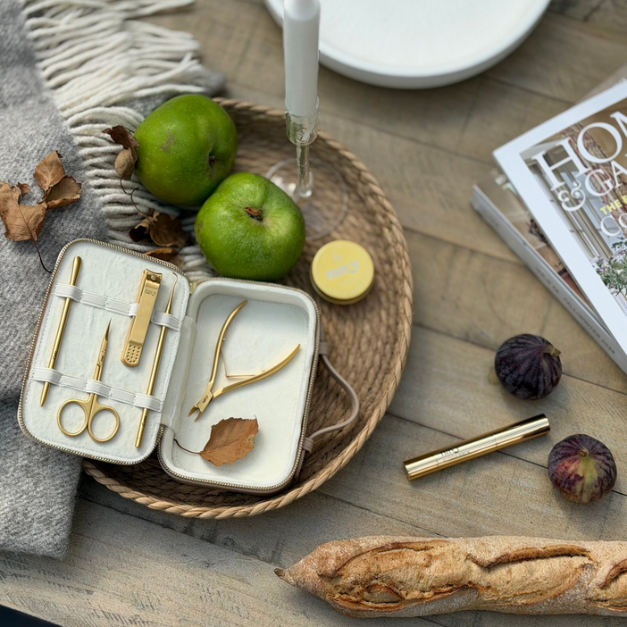 An open metal travel vanity case with gold-tone tools and a white interior sits on a woven tray beside green apples, a candle, a magazine, and a baguette on a rustic wooden table.