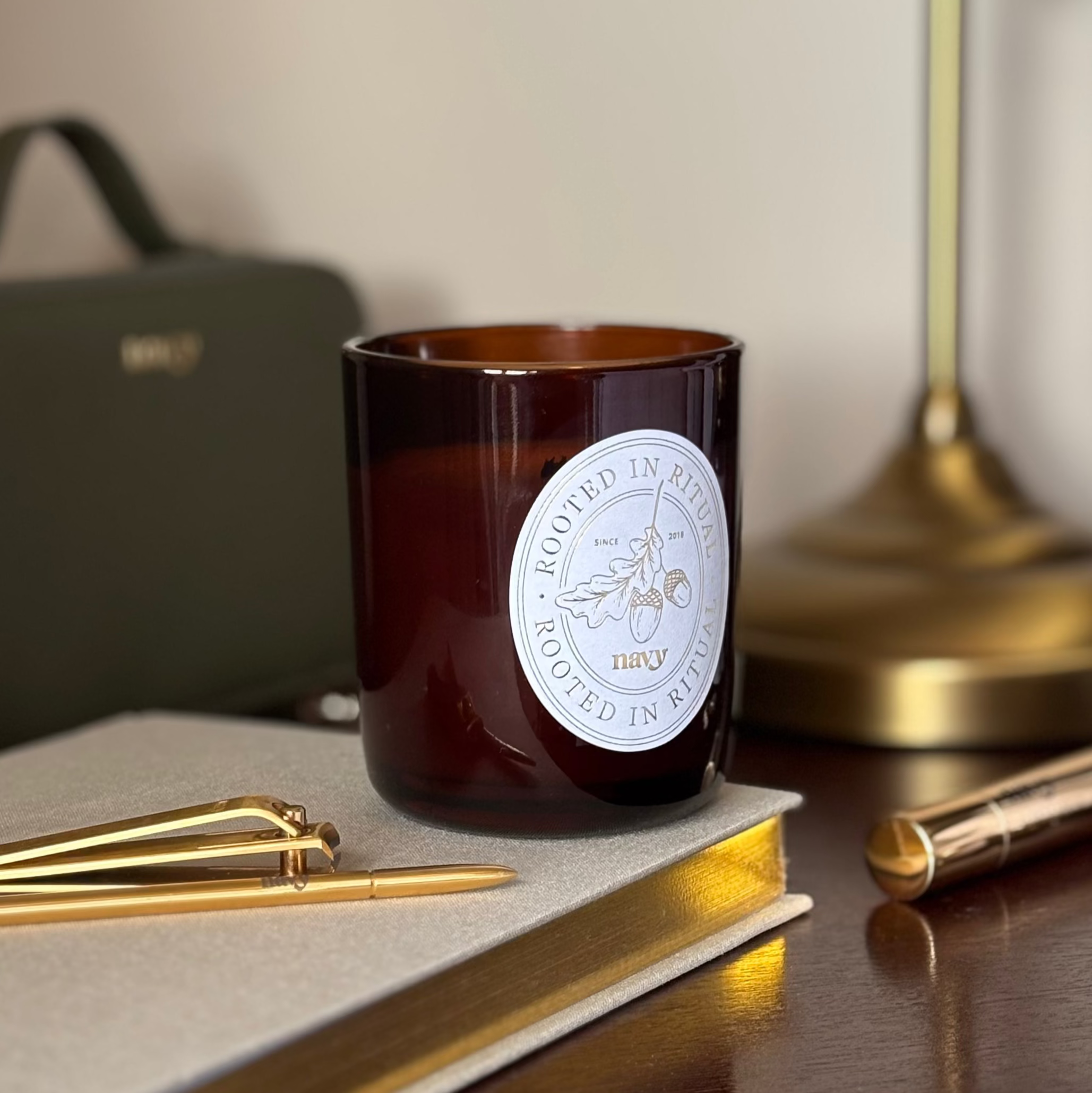 Brown candle with a label on a surface with gold pens and a notebook.