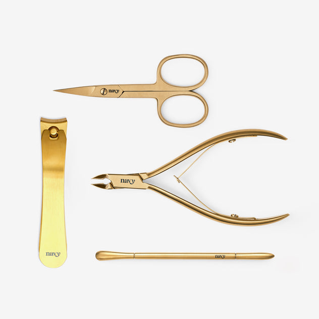 A collection of four gold-colored nail tools including toe nail nips, a curved pusher tool, fine cuticle nips, and curved scissors, all laid out on a white background.