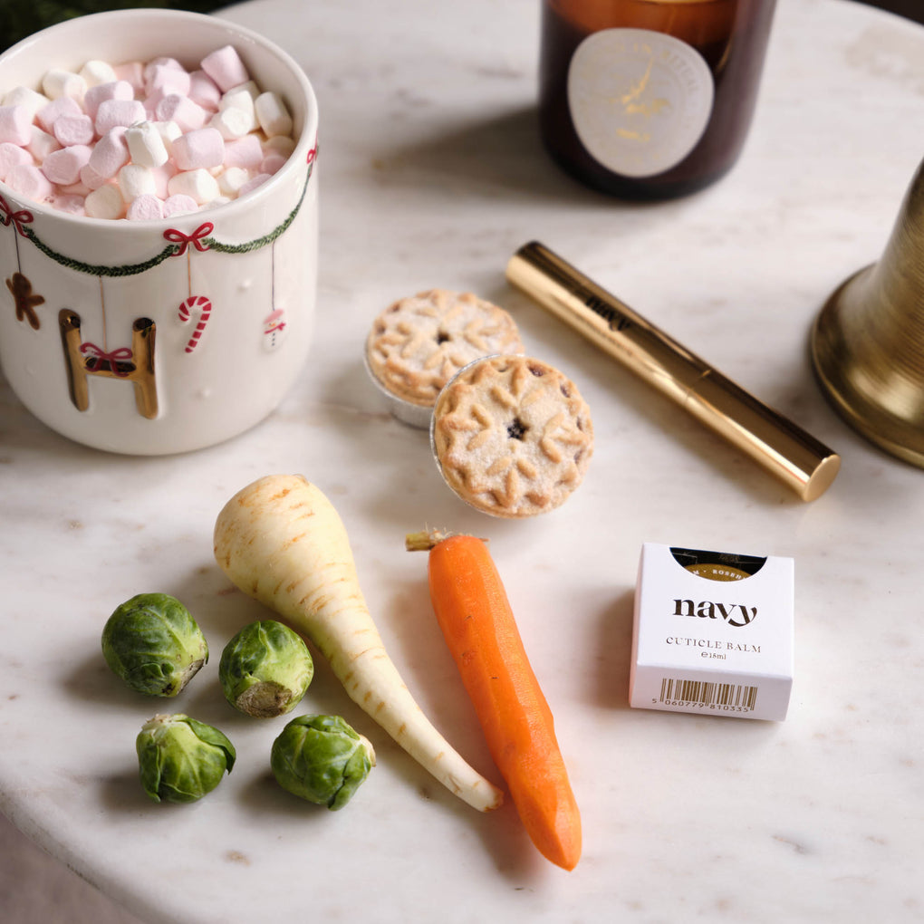 Cup of hot chocolate with marshmallows, cookies, vegetables, and a box of navy cuticle balm on a marble surface.