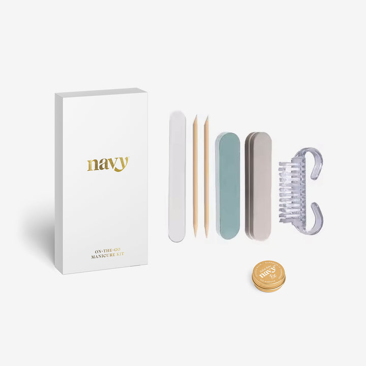 A collection of nail care tools including three nail files, a cuticle balm tin, orange wood sticks, and a reusable nail brush, displayed in front of a white box with the brand name 'navy'.